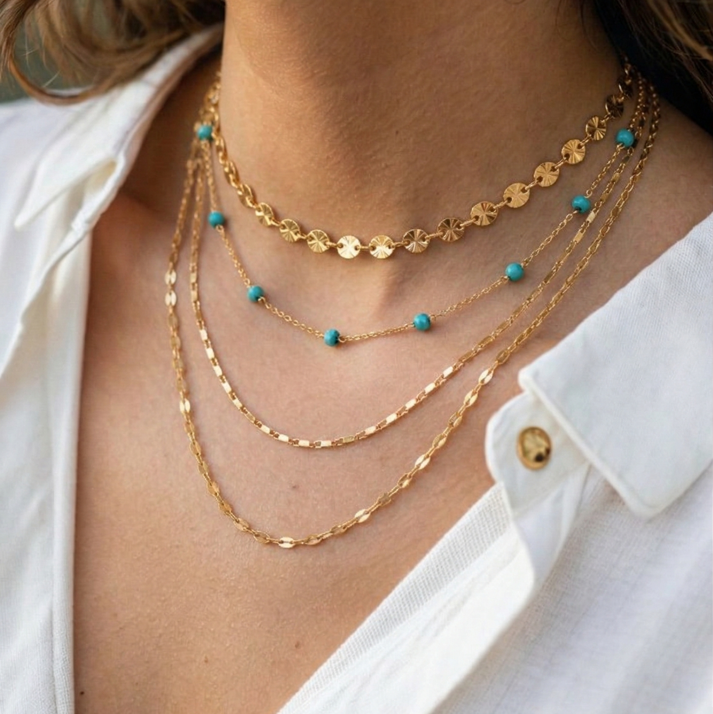 Close-up of a person wearing 4 stacked gold necklaces with turquoise beads on a white shirt.