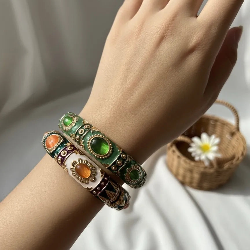 Hand wearing a decorative kada with green and orange stones on a neutral background